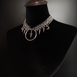 May include: Silver-toned chainmail choker necklace with a gothic aesthetic. The necklace features a large ring pendant with a cross, and is adorned with spikes and crescent moon charms. The jewelry is displayed on a black mannequin.