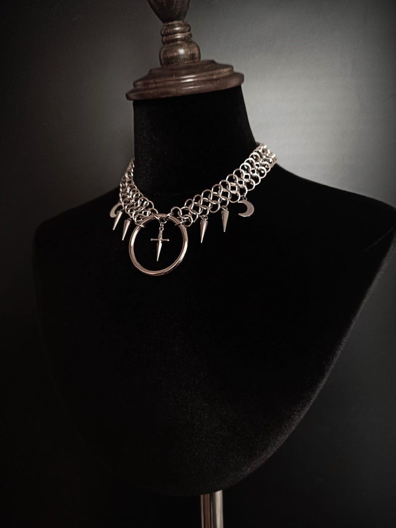 May include: Silver-toned chainmail choker necklace with a gothic aesthetic. The necklace features a large ring pendant with a cross, and is adorned with spikes and crescent moon charms. The jewelry is displayed on a black mannequin.