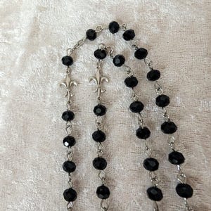 May include: A black beaded rosary with silver metal accents. The beads are round and have a faceted finish. The accents are fleur-de-lis.