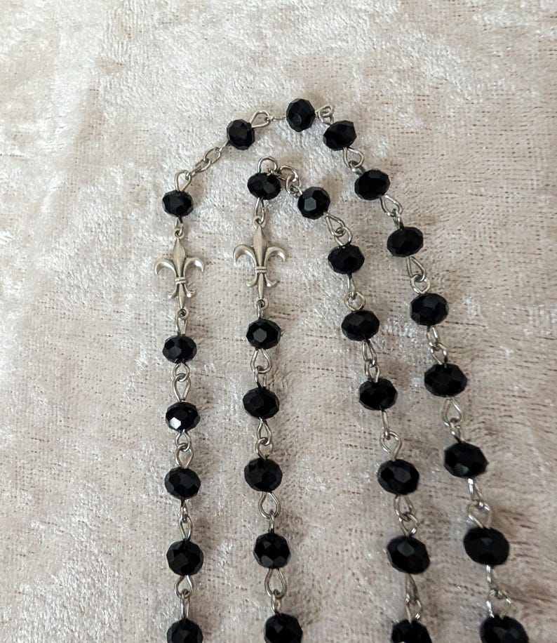 May include: A black beaded rosary with silver metal accents. The beads are round and have a faceted finish. The accents are fleur-de-lis.