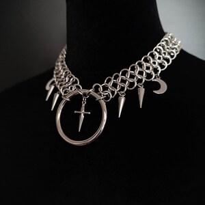 May include: Silver-toned choker necklace with a gothic aesthetic. The necklace features a chainmail-style collar, a large circular pendant with a sword, and dangling charms including spikes and a crescent moon. The necklace is displayed on a black mannequin.