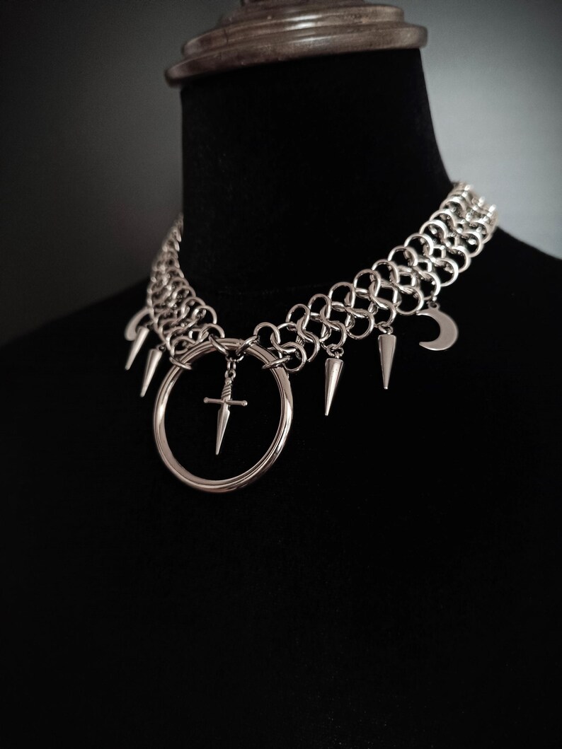 May include: Silver-toned choker necklace with a gothic aesthetic. The necklace features a chainmail-style collar, a large circular pendant with a sword, and dangling charms including spikes and a crescent moon. The necklace is displayed on a black mannequin.