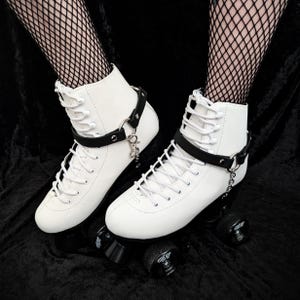 May include: Pair of white roller skates with black wheels and accents. The skates have a high-top design with white laces and a black strap around the ankle. A silver chain hangs from the strap. The skates are on a black surface.