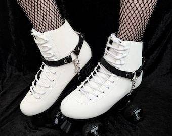 May include: Pair of white roller skates with black wheels and accents. The skates have a high-top design with white laces and a black strap around the ankle. A silver chain hangs from the strap. The skates are on a black surface.