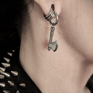 May include: A silver-colored earring set. The earring features a small axe pendant hanging from a hoop earring. The earring is worn in a pierced ear. The image is a close-up shot.
