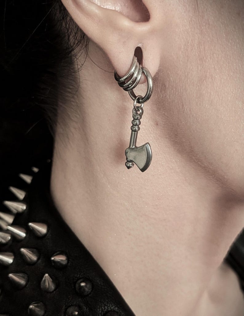 May include: A silver-colored earring set. The earring features a small axe pendant hanging from a hoop earring. The earring is worn in a pierced ear. The image is a close-up shot.