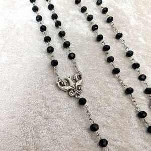 May include: A black beaded rosary necklace with a silver ornate pendant. The beads are faceted and the chain is silver.