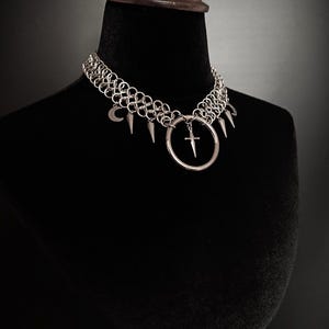 May include: A silver chainmail choker necklace with a gothic aesthetic. The necklace features a series of metal rings, adorned with a crescent moon, spikes, and a large ring with a cross pendant. The necklace is displayed on a black mannequin.