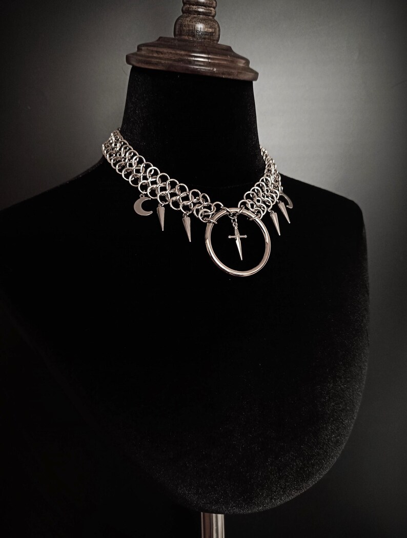 May include: A silver chainmail choker necklace with a gothic aesthetic. The necklace features a series of metal rings, adorned with a crescent moon, spikes, and a large ring with a cross pendant. The necklace is displayed on a black mannequin.