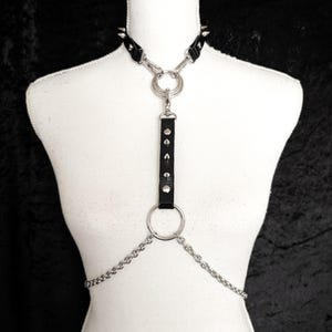 Puede incluir: Un collar de cuero negro con detalles metálicos plateados y una cadena unida a un anillo plateado grande. La cadena se extiende hasta un anillo plateado más pequeño.