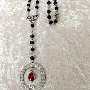 May include: A black beaded necklace with a silver pendant featuring a snake coiled around a red gemstone. The pendant is attached to a silver chain with a decorative clasp.