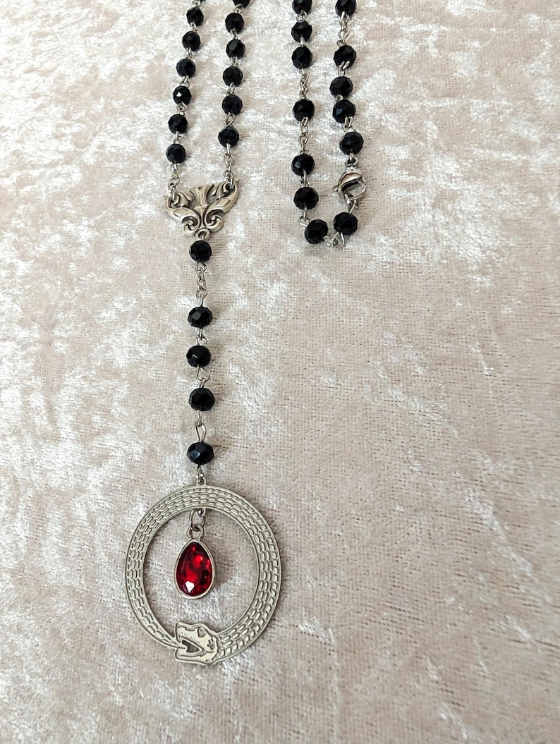 May include: A black beaded necklace with a silver pendant featuring a snake coiled around a red gemstone. The pendant is attached to a silver chain with a decorative clasp.