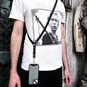 May include: A black leather phone harness with a silver clasp holding a gray phone case. The harness is worn over a white t-shirt with a black and white graphic and the word "GUMMO". A silver chain necklace is also visible.