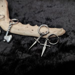 May include: A pair of silver-colored axe-shaped keychains and two pairs of silver dagger earrings with hoop closures. The items are displayed on a dark, textured surface with a piece of light-colored wood.