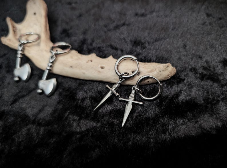 May include: A pair of silver-colored axe-shaped keychains and two pairs of silver dagger earrings with hoop closures. The items are displayed on a dark, textured surface with a piece of light-colored wood.