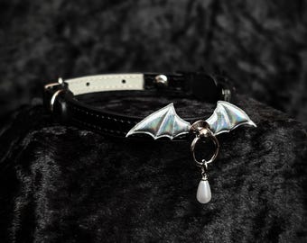 May include: Black leather collar with a silver bat wing charm and a pearlescent teardrop pendant. The collar has a silver metal ring and buckle. The inside of the collar is white. The bat wings have a holographic sheen.