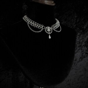 K&ouml;nnte beinhalten: Ein silberfarbenes Choker-Halsband mit filigranem Design. Die Halskette hat mehrere verbundene Ringe, einen herzf&ouml;rmigen Mittelanh&auml;nger mit einem kleinen Edelstein und eine tropfenf&ouml;rmige Perle. Pr&auml;sentiert auf einer schwarzen Schaufensterpuppe.