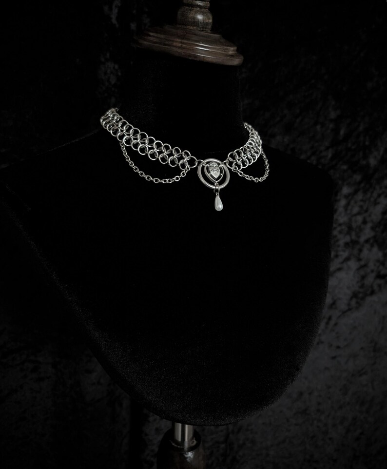K&ouml;nnte beinhalten: Ein silberfarbenes Choker-Halsband mit filigranem Design. Die Halskette hat mehrere verbundene Ringe, einen herzf&ouml;rmigen Mittelanh&auml;nger mit einem kleinen Edelstein und eine tropfenf&ouml;rmige Perle. Pr&auml;sentiert auf einer schwarzen Schaufensterpuppe.