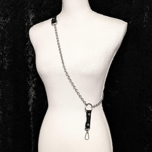 Puede incluir: Un arnés de cadena plateada con una correa de cuero negro y un cierre de metal. La cadena se conecta a un lazo de cuero negro en el hombro y a un anillo de metal en la cintura. El arnés se muestra en un maniquí blanco.