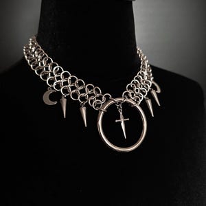 May include: Silver-toned choker necklace with a chainmail design. The necklace features a large ring pendant with a small cross, along with dangling spikes and a crescent moon charm. The necklace is displayed on a black mannequin.