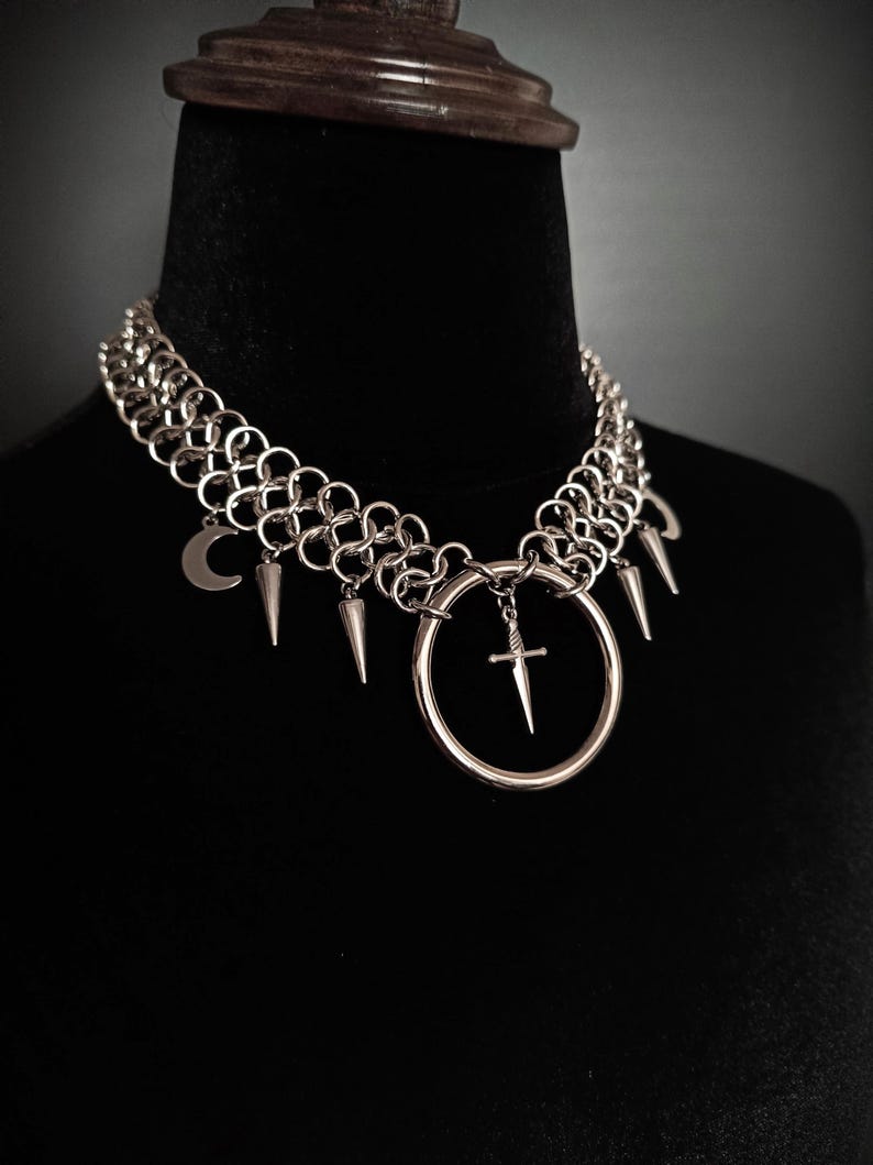 May include: Silver-toned choker necklace with a chainmail design. The necklace features a large ring pendant with a small cross, along with dangling spikes and a crescent moon charm. The necklace is displayed on a black mannequin.