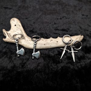 May include: Silver-toned axe and sword earrings displayed on a bone against a black background. The earrings are small, detailed, and have a metallic sheen. The axes and swords are attached to small hoops.