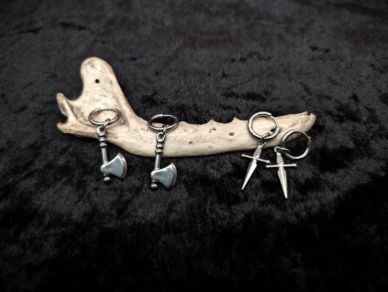 May include: Silver-toned axe and sword earrings displayed on a bone against a black background. The earrings are small, detailed, and have a metallic sheen. The axes and swords are attached to small hoops.