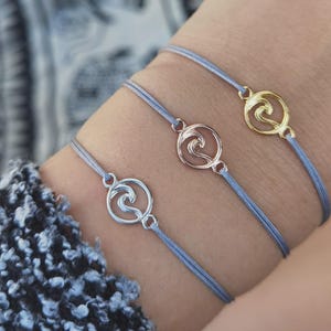 Könnte beinhalten: Drei Armbänder mit Wellenanhängern in Silber, Roségold und Gold. Die Armbänder haben eine hellblaue Kordel. Die Wellenanhänger sind kreisförmig und weisen ein stilisiertes Wellendesign auf.