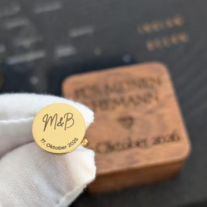 Personalisierbarer Manschettenknopf aus goldfarbenem Edelstahl mit eingravierten Initialen „M & B“ und Hochzeitsdatum, gehalten in weißem Handschuh; ideale Geschenkidee für Bräutigam, Trauzeugen oder Hochzeit, hochwertige Gravur, Holzbox im Hintergrund.