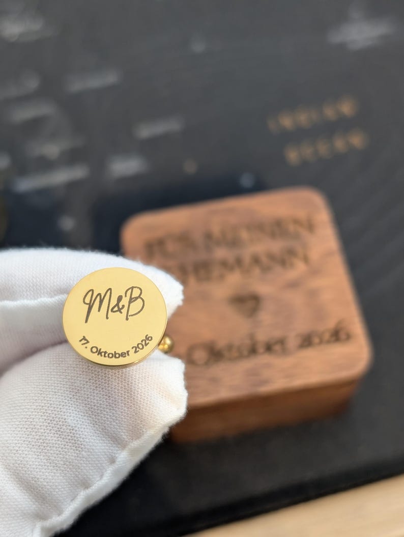 Personalisierbarer Manschettenknopf aus goldfarbenem Edelstahl mit eingravierten Initialen „M & B“ und Hochzeitsdatum, gehalten in weißem Handschuh; ideale Geschenkidee für Bräutigam, Trauzeugen oder Hochzeit, hochwertige Gravur, Holzbox im Hintergrund.