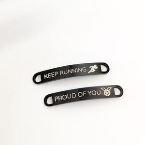 Puede incluir: Dos etiquetas de zapatos de metal negro con texto blanco. Una etiqueta dice "KEEP RUNNING" con un icono de corredor. La otra etiqueta dice "PROUD OF YOU" con un icono de medalla. Ambas etiquetas tienen forma curva con un agujero en cada extremo.