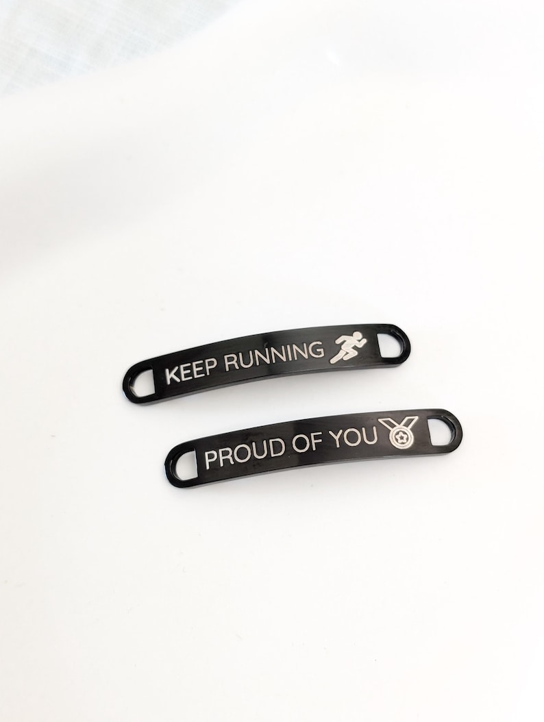 Puede incluir: Dos etiquetas de zapatos de metal negro con texto blanco. Una etiqueta dice "KEEP RUNNING" con un icono de corredor. La otra etiqueta dice "PROUD OF YOU" con un icono de medalla. Ambas etiquetas tienen forma curva con un agujero en cada extremo.