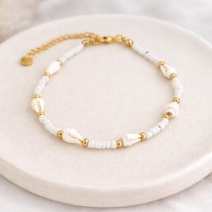 Può includere: Una delicata cavigliera con perline bianche, piccole conchiglie e dettagli color oro. La cavigliera è esposta su una superficie bianca, mostrando il suo design intricato e l'estetica da spiaggia. La catena è regolabile.