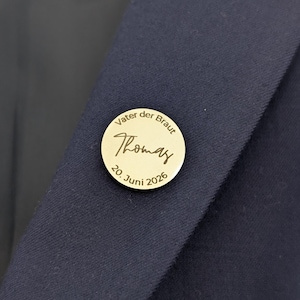 Puede incluir: Pin redondo dorado con la inscripción "Vater der Braut Thomas 20. Juni 2026". El pin está sujeto a una tela azul oscuro, probablemente una chaqueta de traje. Un accesorio de boda.