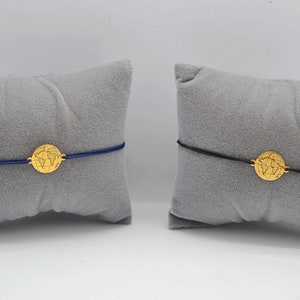 Zwei goldene Weltkarte Armbänder auf grauen Schmuckkissen. Ein Armband mit blauem Band und eins mit schwarzem Band