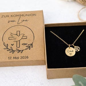 Puede incluir: Un collar dorado con dos colgantes: uno redondo con el nombre "Lina" y un corazón, y un árbol de la vida. Presentado en una caja negra, junto a una caja marrón con texto y un diseño de cruz. La fecha "17. Mai 2026" es visible.