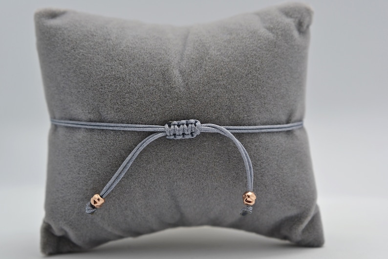 Makramee Verschluss Armband graues Band mit roségoldenen Perlen auf einem grauen Schmuckkissen