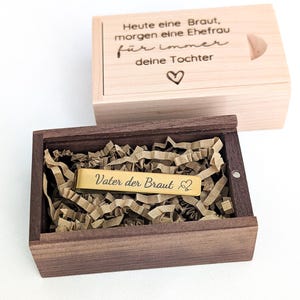Può includere: Una scatola di legno con coperchio incernierato, contenente una barra color oro con la scritta "Vater der Braut" e un simbolo a forma di cuore. Il coperchio della scatola sopra ha la scritta "Heute eine Braut, morgen eine Ehefrau für immer deine Tochter" e un simbolo a forma di cuore.