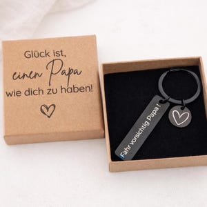 Könnte beinhalten: Ein schwarzer Schlüsselanhänger mit einem Herzanhänger und dem Text "Fahr vorsichtig Papa!" wird in einer braunen Schachtel neben einer Schachtel mit dem Text "Glück ist, einen Papa wie dich zu haben!" und einem Herzsymbol angezeigt.