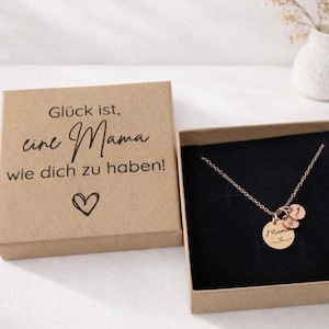 Puede incluir: Un collar de oro rosa con tres pequeños colgantes circulares grabados, presentado en una caja de regalo de cartón marrón. La tapa de la caja tiene impreso el texto en alemán 'Glück ist, eine Mama wie dich zu haben!' y un corazón.
