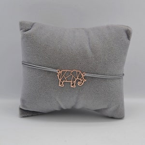 Könnte beinhalten: Ein Armband mit einem geometrischen Elefanten-Charm aus Roségold. Der Charm ist an einer grauen Kordel befestigt. Das Armband wird auf einem grauen Samtkissen vor weißem Hintergrund präsentiert. Der Elefanten-Charm ist ca. 2,5 cm lang.