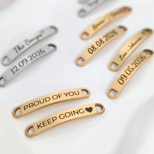 Könnte beinhalten: Nahaufnahme von mehreren gebogenen Metallanhängern. Einige sind silberfarben, andere goldfarben. Die Anhänger tragen eingravierte Texte, darunter Namen, Daten und Sätze wie "PROUD OF YOU" und "KEEP GOING".