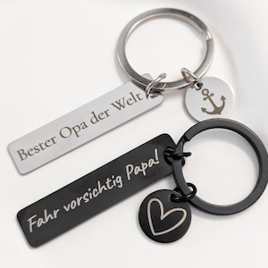 Könnte beinhalten: Zwei Schlüsselanhänger mit silbernen und schwarzen Metallringen. Ein silberner Schlüsselanhänger trägt den Text "Bester Opa der Welt". Der schwarze Schlüsselanhänger trägt den Text "Fahr vorsichtig Papa!" und einen Herzanhänger. Ein Ankeranhänger ist ebenfalls vorhanden.