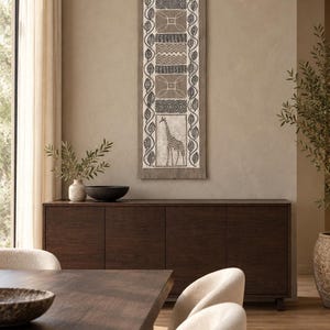 May include: A vertical wall hanging featuring a rectangular fabric panel with a dark brown wooden top bar. The design includes two giraffe illustrations at the top and bottom, separated by three central panels with geometric patterns and leaf borders.