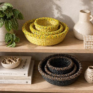 May include: A wooden shelf displays decorative items. Three yellow woven baskets are stacked together. A potted plant, a white vase, a small patterned box, and two dark blue woven baskets are also present. A small, decorative vase and books complete the display.