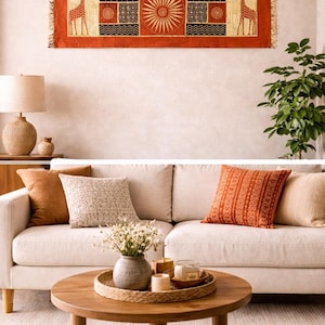 May include: A living room scene featuring a sofa with decorative pillows, a round wooden coffee table, and a wall hanging. The wall hanging is rectangular with a red background and giraffe and sun designs. A lamp and plant add to the decor.