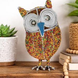 Może przedstawiać: Kolorowa figurka sowy z koralików. Sowa ma duże oczy owinięte srebrnym drutem i niebieski dziób. Ciało pokryte jest mozaiką z czerwonych, żółtych i zielonych koralików. Sowa stoi na drewnianej półce.