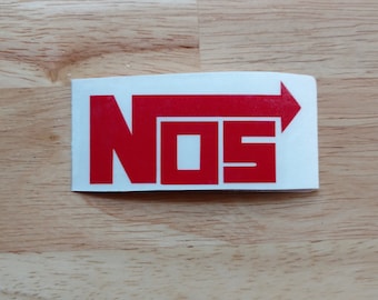 May include: A rectangular white sticker with the red letters "NOS" and an arrow pointing right. The sticker is on a light brown wooden surface.