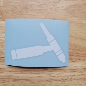 May include: A white vinyl sticker featuring a simplified illustration of a welding torch on a light blue background. The sticker is rectangular with rounded corners and is placed on a wooden surface.
