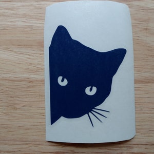 May include: A navy blue cat silhouette peeking from the side, with white eyes and whiskers, on a white sticker. The sticker is placed on a light brown wooden surface.
